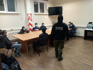 Kobieta i 6mężczyzn siedzi w maseczkach chirurgicznych przy stole, obok stoi funckjoanriusz Straży Granicznej