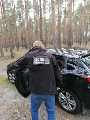 zatrzymany Mołdawianin siedzi skuty kajdankami na tylnym siedzeniu granatowego auta obok stoi funckjoanriusz Straży Granicznej