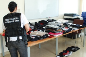 Zatrzymane podrobione ubrania na stole leżą podrobione ubrania różnych znanych światowych marek, obok stoi funkcjonariuszka SG po cywilnemu, na sobie ma kamizelkę taktyczną koloru czarnego z napisem Straż Graniczna