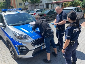 Sprawca kradzieży rozbójniczej zatrzymany na gorącym uczynku przez funkcjonariusza Straży Granicznej sprawca kradzieży stoi oparty przodem rękoma o policyjny radiowóz, obok niego stoi policjantka i policjant w mundurach służbowych